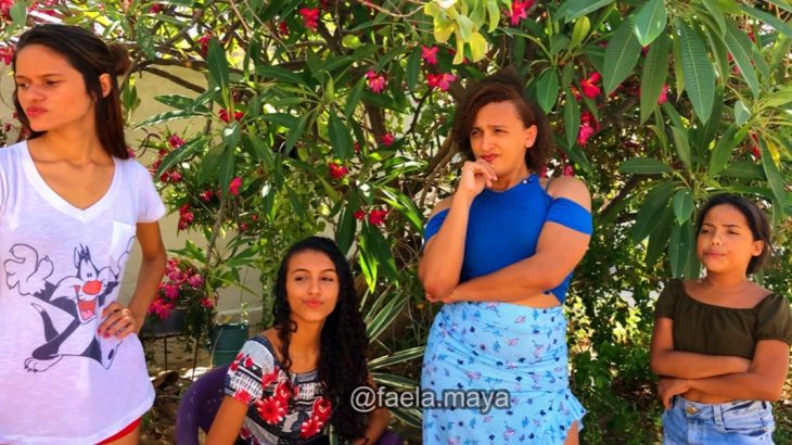 Cena de vídeo do canal de Faela Maya: três mulheres e uma garota debaixo de uma árvore florida na rua, olhando com desprezo e deboche para o canto esquerdo do quadro