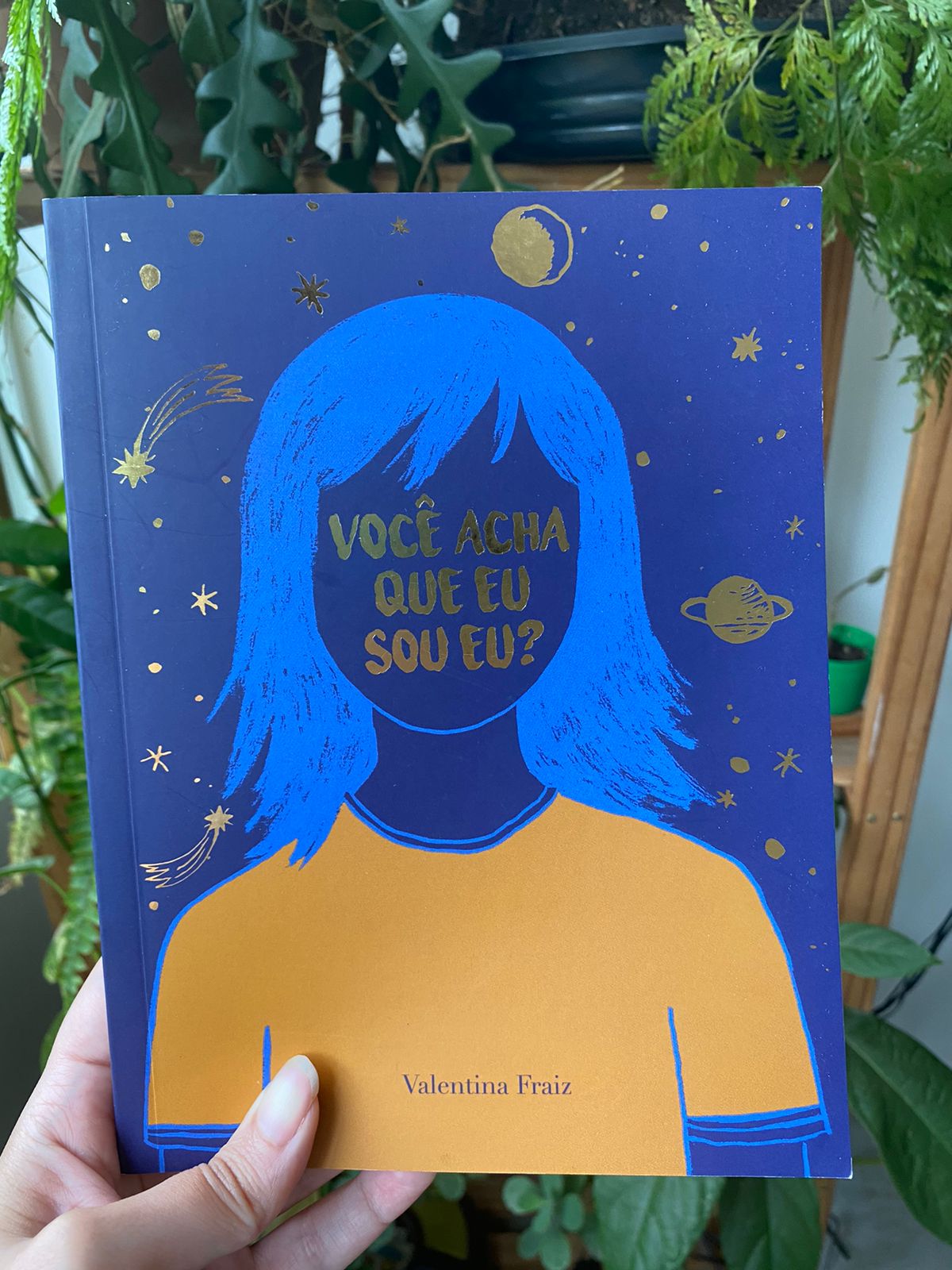 Foto da capa do livro: Você acha que eu sou eu? O título é escrito em dourado no lugar do rosto de uma garota com cabelos azuis, cercada de planetas e estrelas também dourados