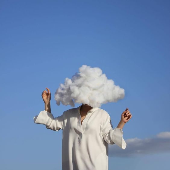 Fotografia de uma mulher com uma nuvem no lugar de sua cabeça. Ela usa uma camisa social branca com as mangas desabotoadas, os braços levemente erguidos, posando sobre um céu absolutamente azul ao fundo.