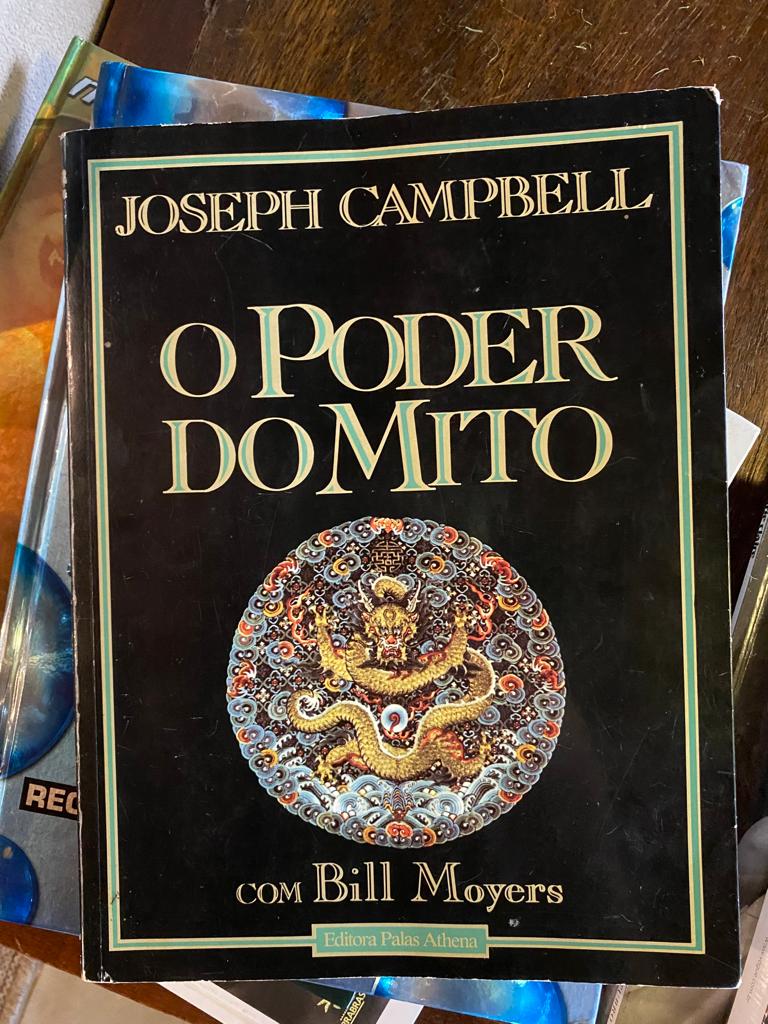 Foto da capa do livro O Poder do Mito de Joseph Campbell com Bill Moyers, edição da Editora Palas Athena. A capa é preta, com fontes serifadas em caixa alta. No centro, um símbolo com um dragão dourado cercado de padrões redondos, nas cores vermelha e azul.