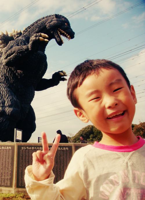 Fotografia de um garotinho asiático sorrindo para a câmera e fazendo "V" com os dedinhos, posando diante de um enorme Godzilla ao fundo.